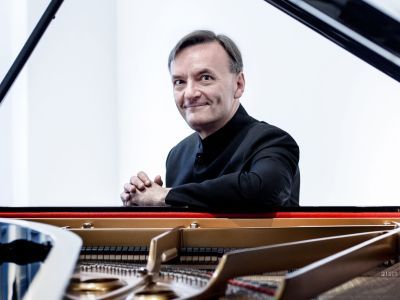 A smiling Stephen Hough wearing a black suit sat behind a piano with a white backdrop